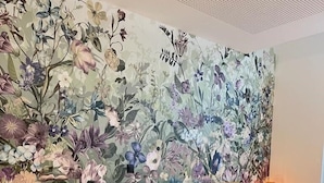 Un dormitorio con papel tapiz floral, cabecera de madera, dos mesitas de noche con lámparas y un escritorio con lámpara.