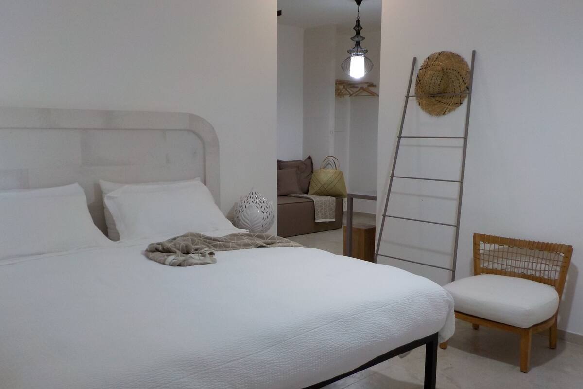 A neatly made bed with white linens, a wooden chair, a ladder, and a woven basket on the wall.