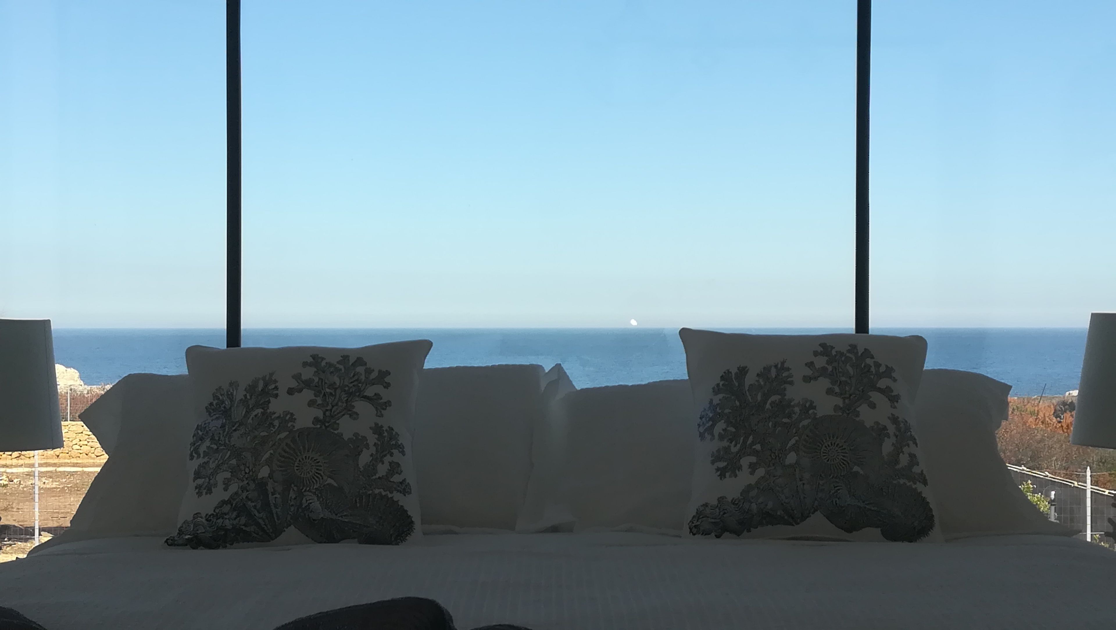 A bed with two decorative pillows and a view of the sea through large windows.