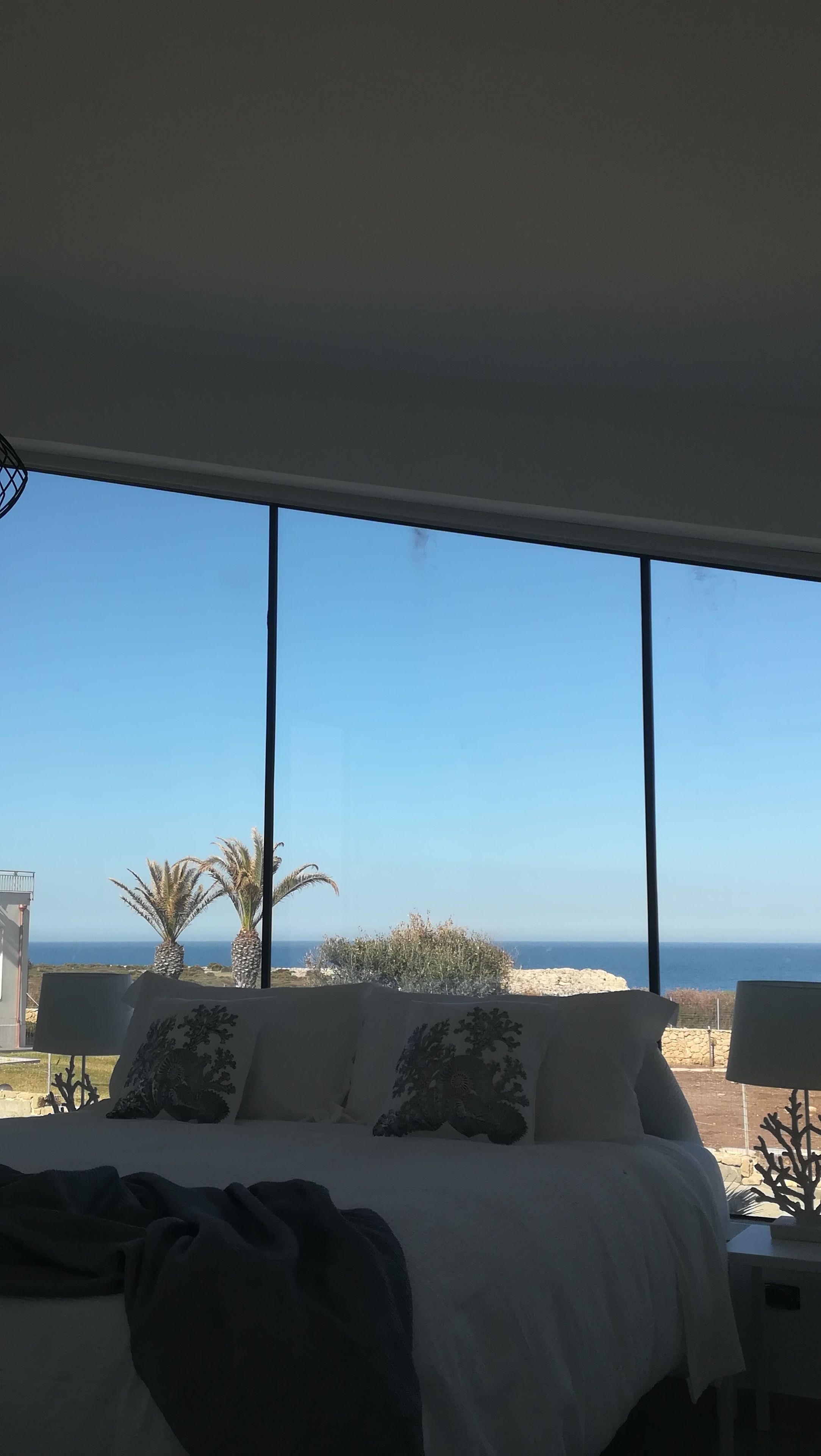 Un dormitorio con un ventanal grande con vistas a una playa y al océano. Hay una cama con ropa de cama blanca y almohadas decorativas.