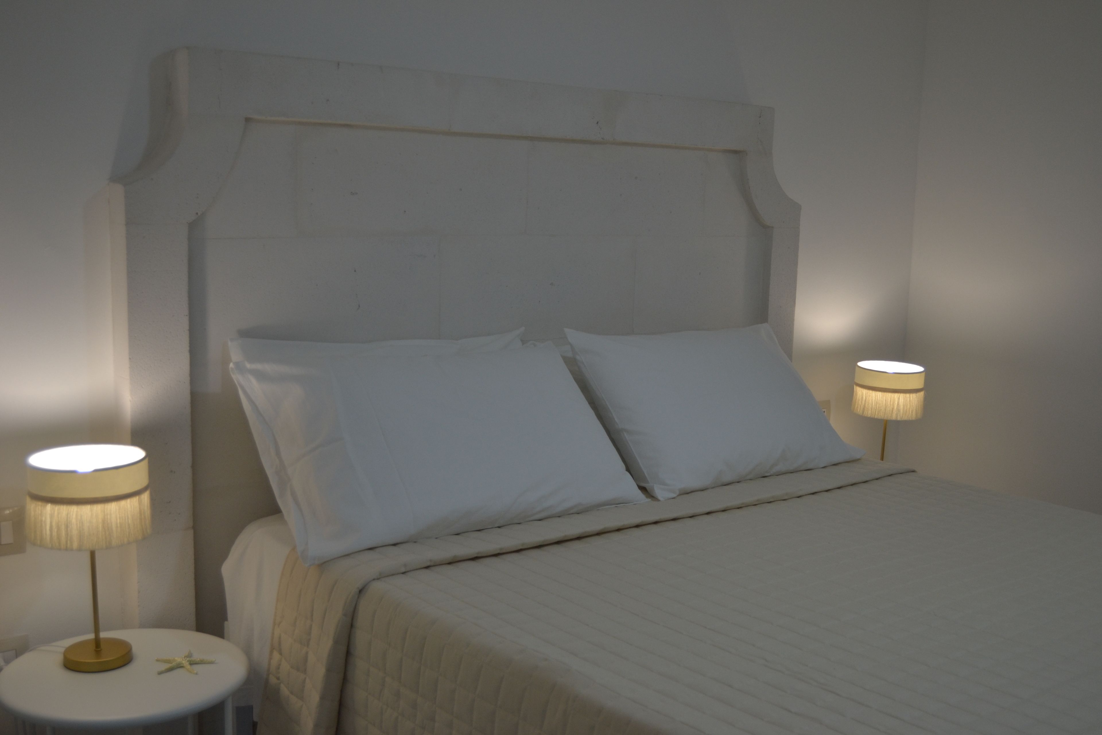 Una cama bien tendida con un cabecero, dos lámparas de noche y una mesita redonda con una lámpara.