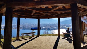 A wooden pavilion with a bench and a table, overlooking a lake and mountains. - Ferienwohnung Landhaus alpinum (Lenggries)