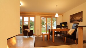 A cozy living room with a fireplace, a dining table with chairs, and a large window offering a view of the outdoors. - Ferienwohnung Landhaus alpinum (Lenggries)