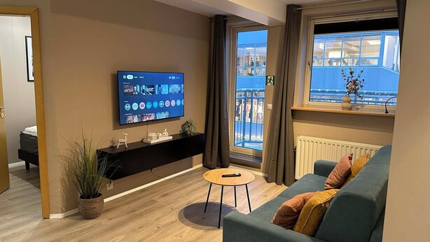 A modern living room with a flat-screen TV, a sofa, a small table, and a window with a view of a building. - Saga Apartments (Akureyri)