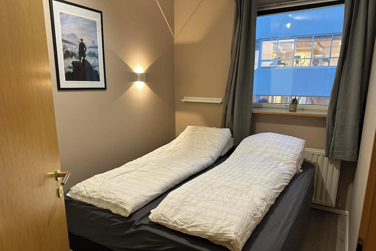 A small room with two single beds, a framed picture on the wall, a window with curtains, and a radiator.