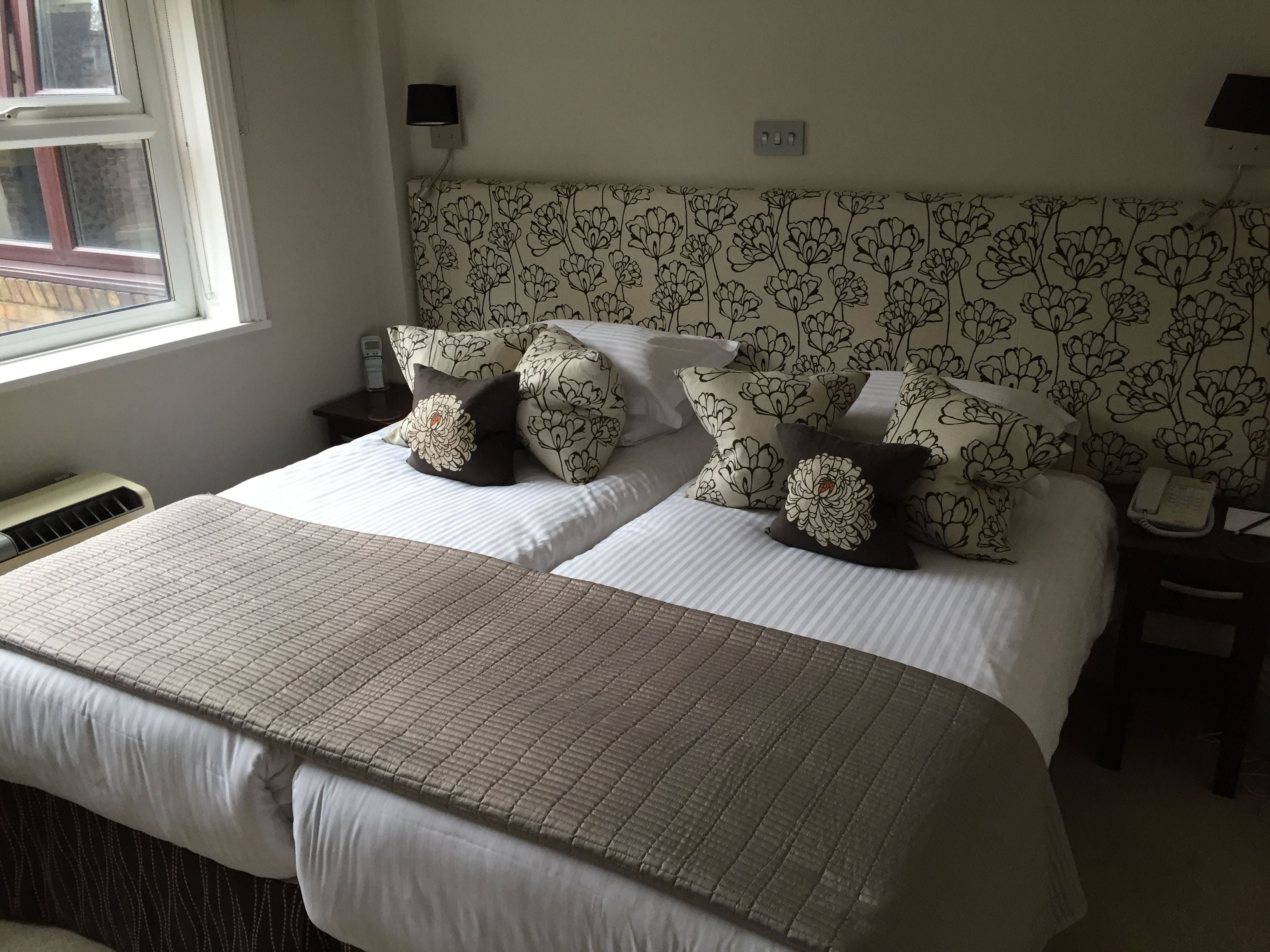 Ein Doppelbett mit einem gemusterten Kopfteil, zwei Nachttische mit Lampen, ein Nachttischtelefon und ein Fenster mit Jalousien.