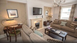 A living room with a fireplace, a television mounted on the wall, a sofa, a coffee table, and a desk with a lamp. - Beaufort House - Knightsbridge (London)