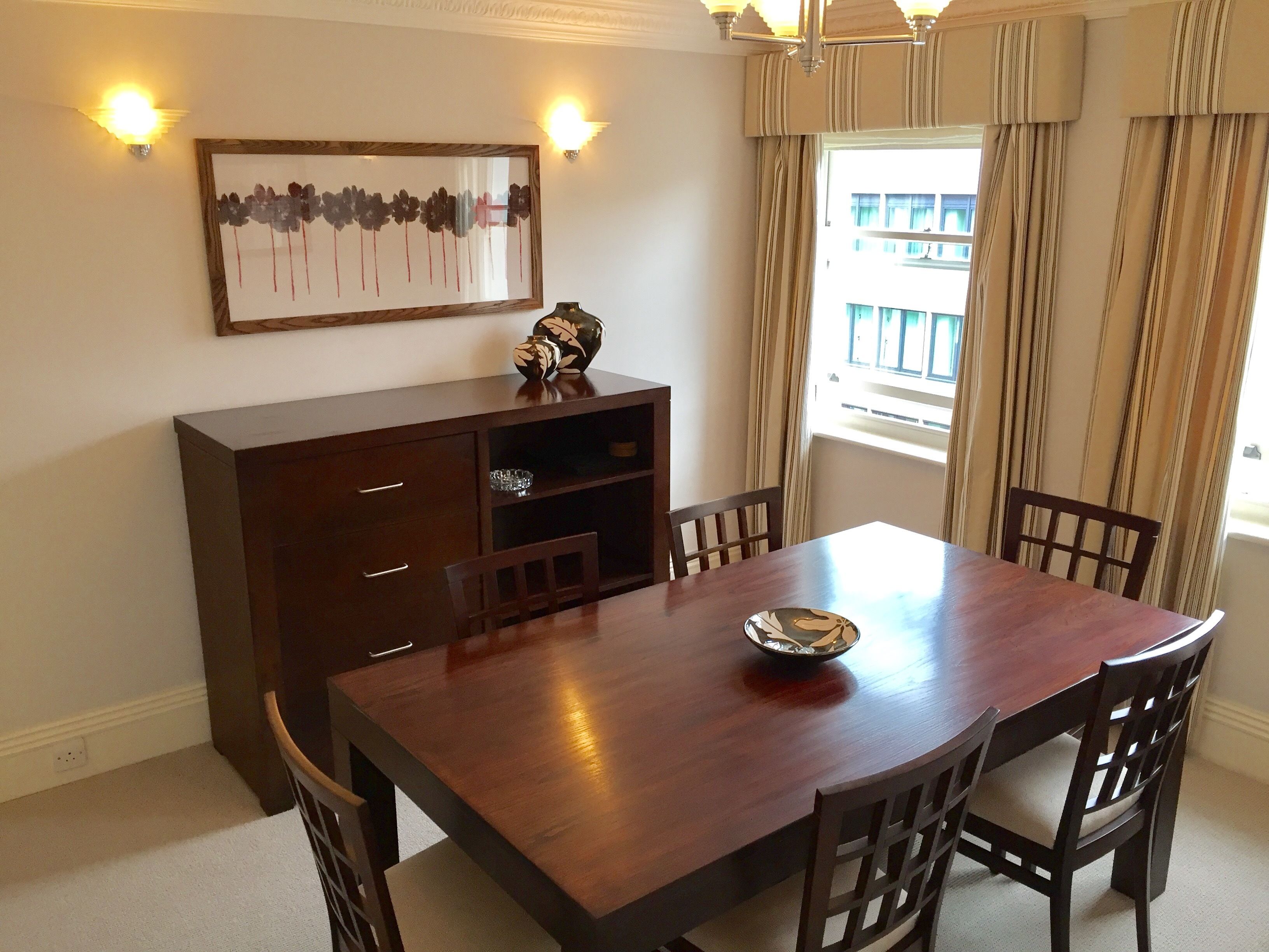 A dining room with a wooden table, six chairs, a decorative vase, and a framed artwork on the wall.