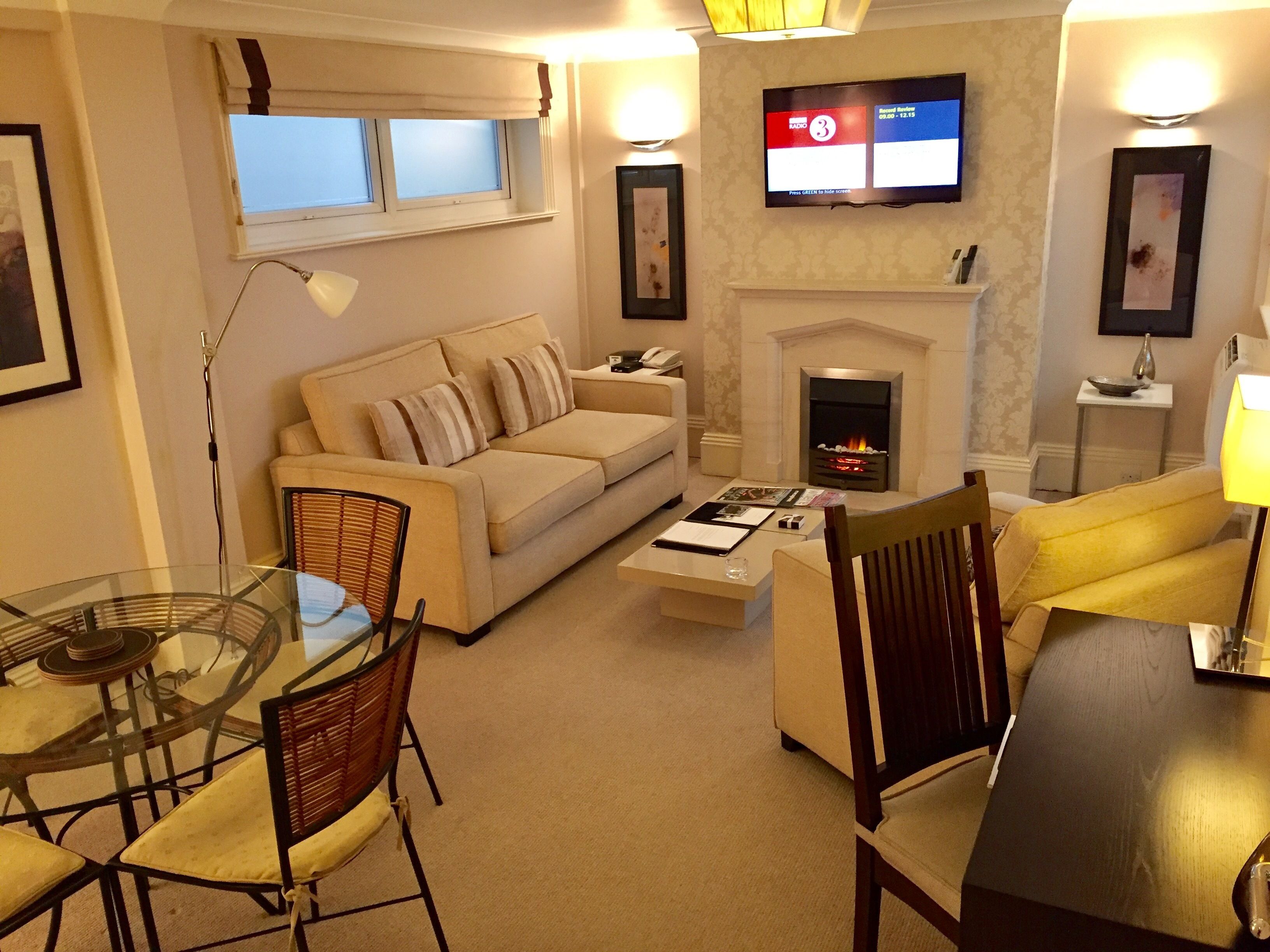 A living room with a sofa, chairs, a glass table, a fireplace, and a TV mounted on the wall.