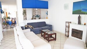 Una sala de estar con sofá, mesa de centro y chimenea. Se observa un comedor con sillas y una mesa al fondo.