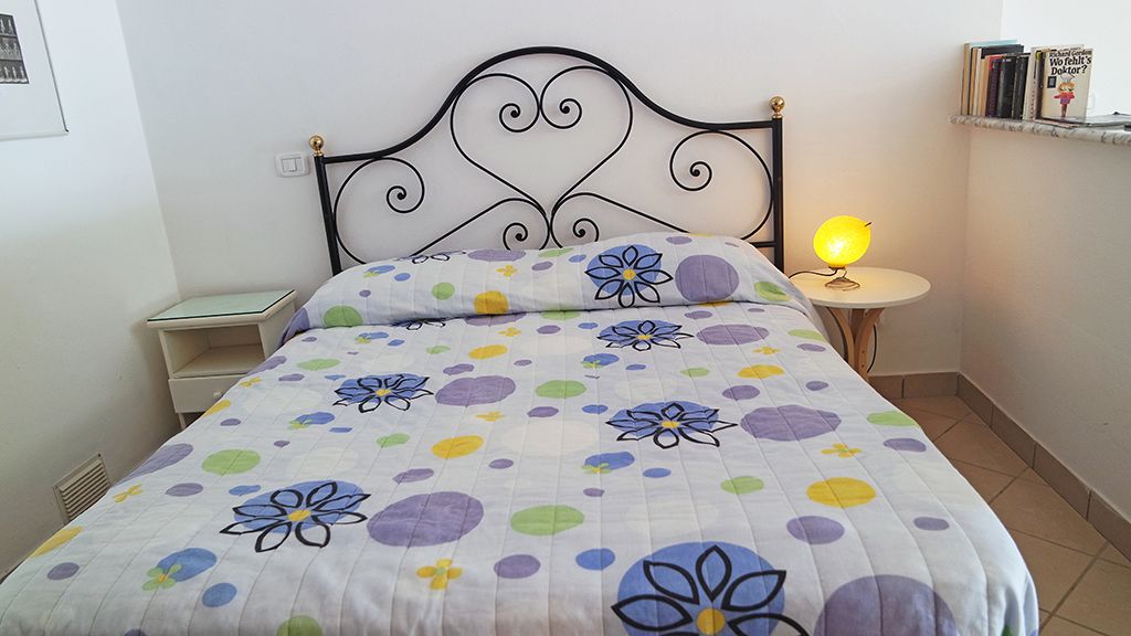 A neatly made bed with a floral-patterned bedspread, a bedside table with a lamp, and a small shelf with books.