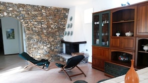 A living room with a stone wall, a fireplace, two lounge chairs, and a wooden cabinet with glass doors. - Villa Sarolli (Ascea)