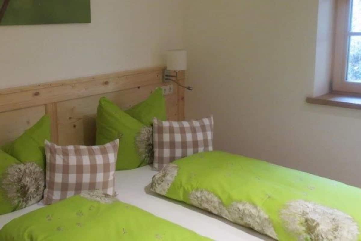 A bedroom with a wooden headboard, two beds with green bedding, a checkered pillow, and a window with daylight.