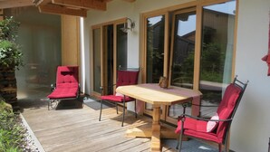 A wooden deck with a table and chairs, a glass door leading to a garden, and a stone wall. - Mountainlodge Dorfhaus (Lenggries)