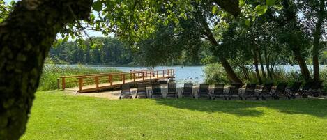 A lakeside park with a wooden deck, lounge chairs, and a grassy area.