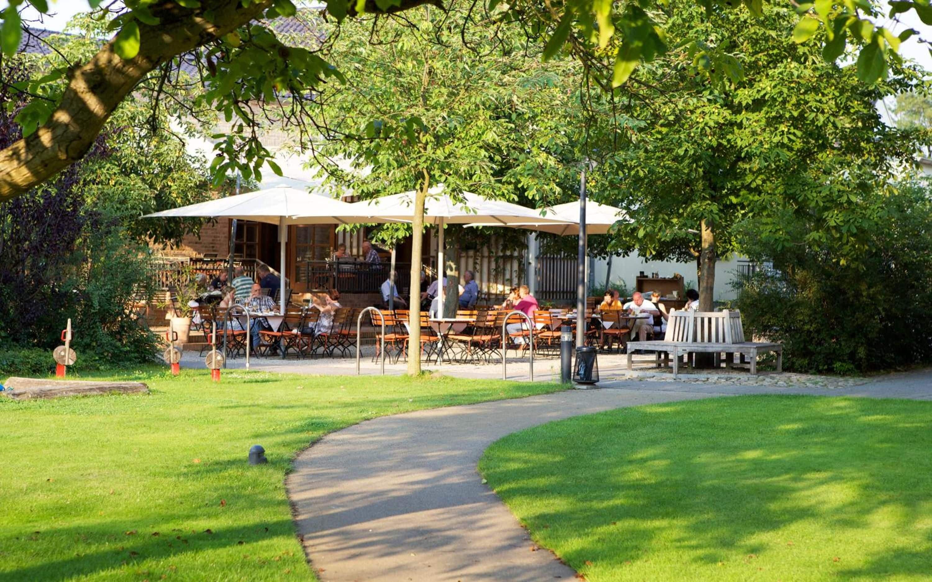 An outdoor seating area with tables and chairs, surrounded by greenery and a pathway.