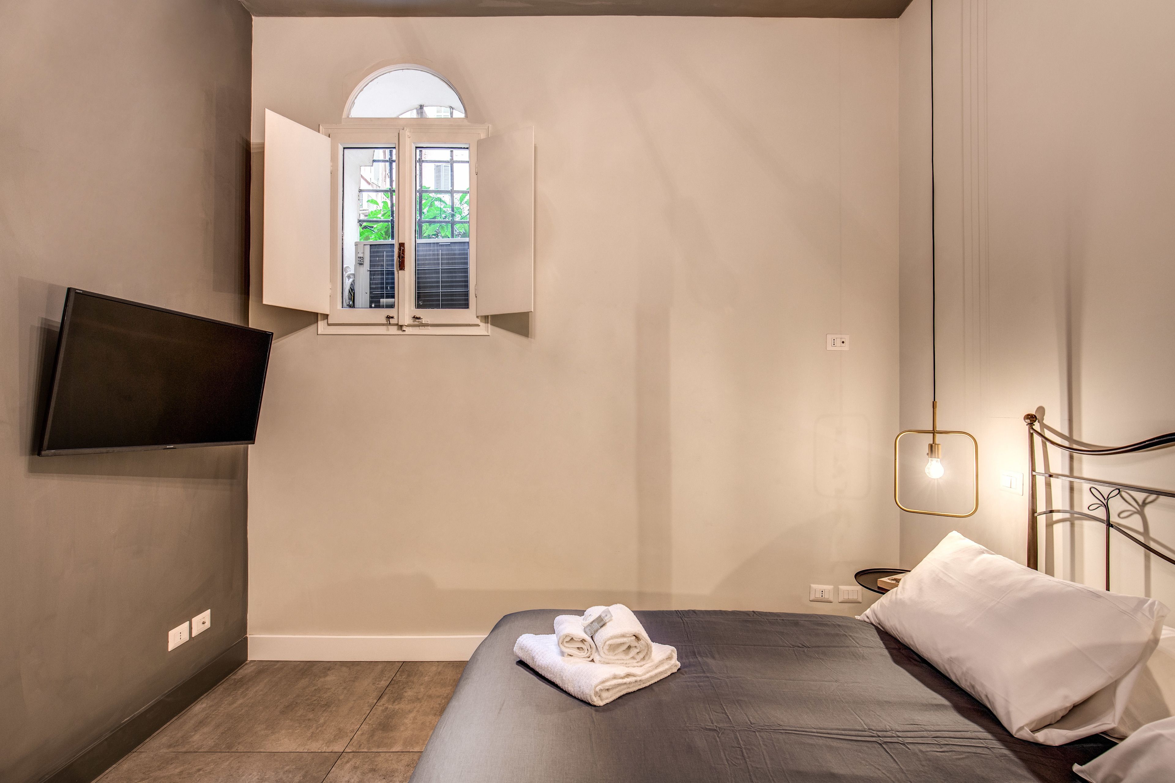 Un dormitorio con una cama, un televisor montado en la pared, una ventana con contraventanas abiertas y una mesita de noche con una lámpara.