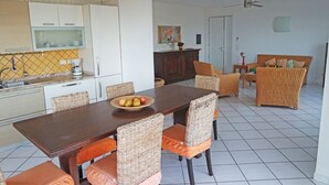 Ein Essbereich mit Holztisch und Korbstühlen, eine Küche mit weißen Schränken sowie ein Wohnbereich mit Sofa und Deckenventilator.