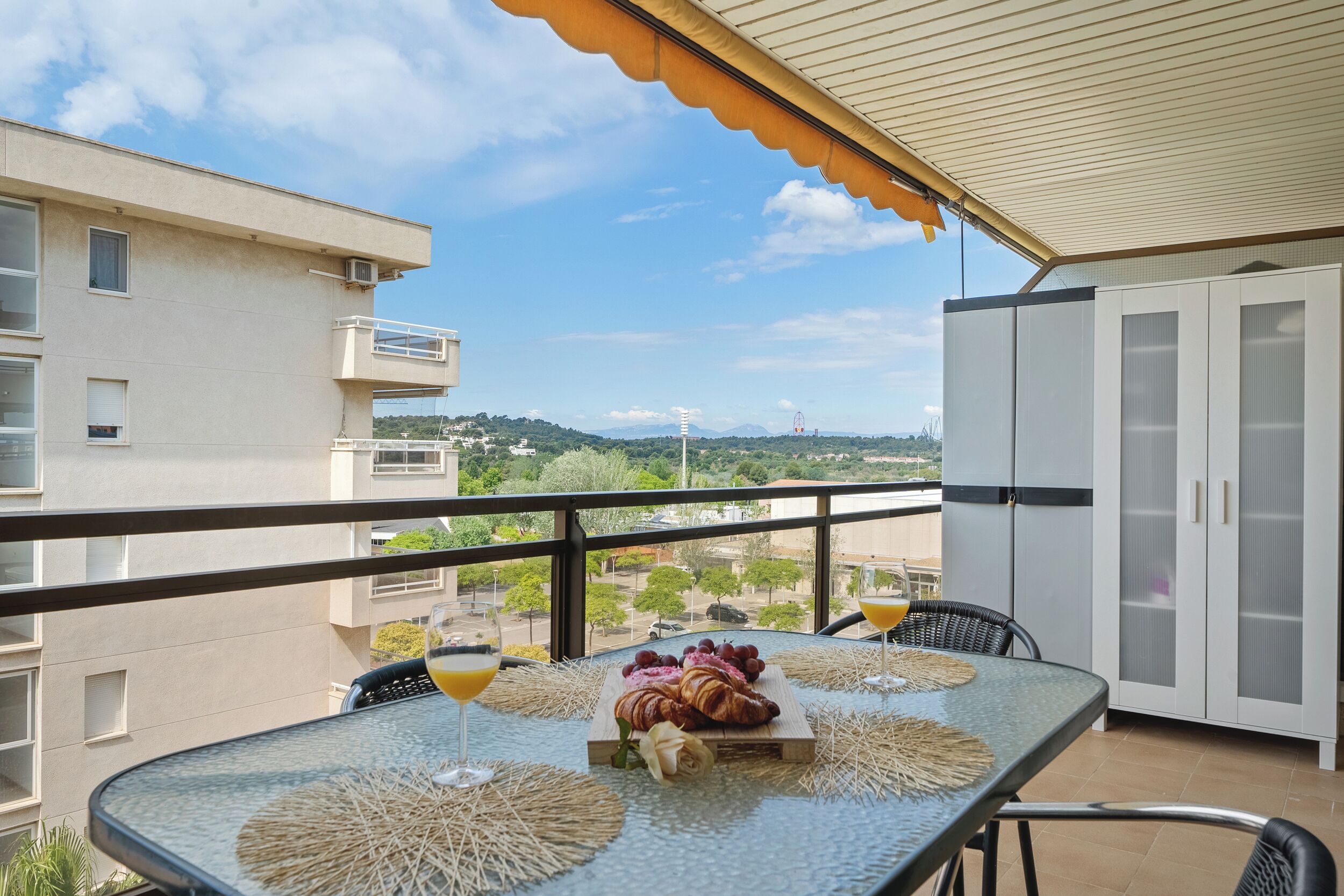 Un balcón con una mesa dispuesta para el desayuno, que incluye croissants y fruta, y vistas de los edificios y el paisaje circundante.