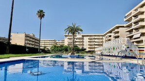 Una piscina con piso de baldosas, rodeada de palmeras y un edificio de varios pisos con balcones.