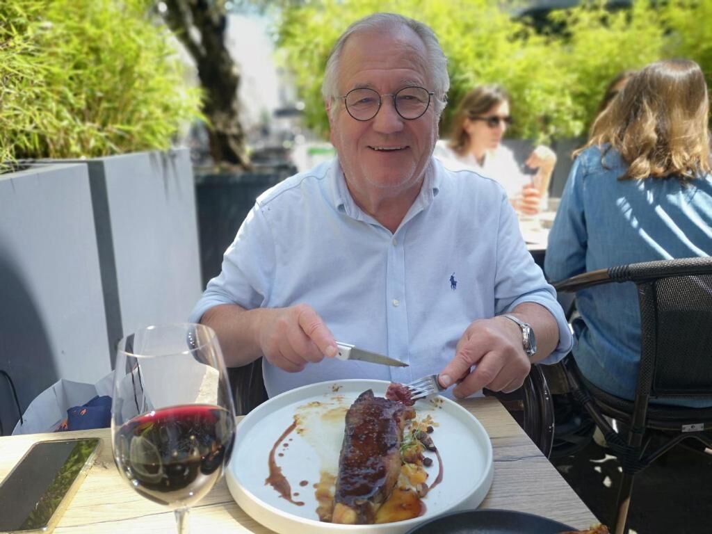 Una persona sentada en una mesa al aire libre, con un plato de comida y una copa de vino tinto.