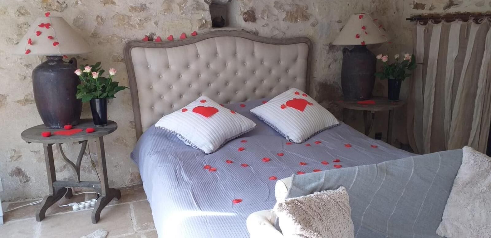 Une chambre à coucher avec une tête de lit capitonnée, deux tables de chevet avec des lampes, un lit recouvert d’une courtepointe bleue et agrémenté d’oreillers à motifs de cœurs, et un vase avec des fleurs sur chaque table.