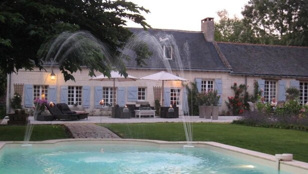 A swimming pool with a fountain, outdoor seating, and a building with shutters. - Chambres d'Hôtes Le Mouton (Blaison-Saint-Sulpice)