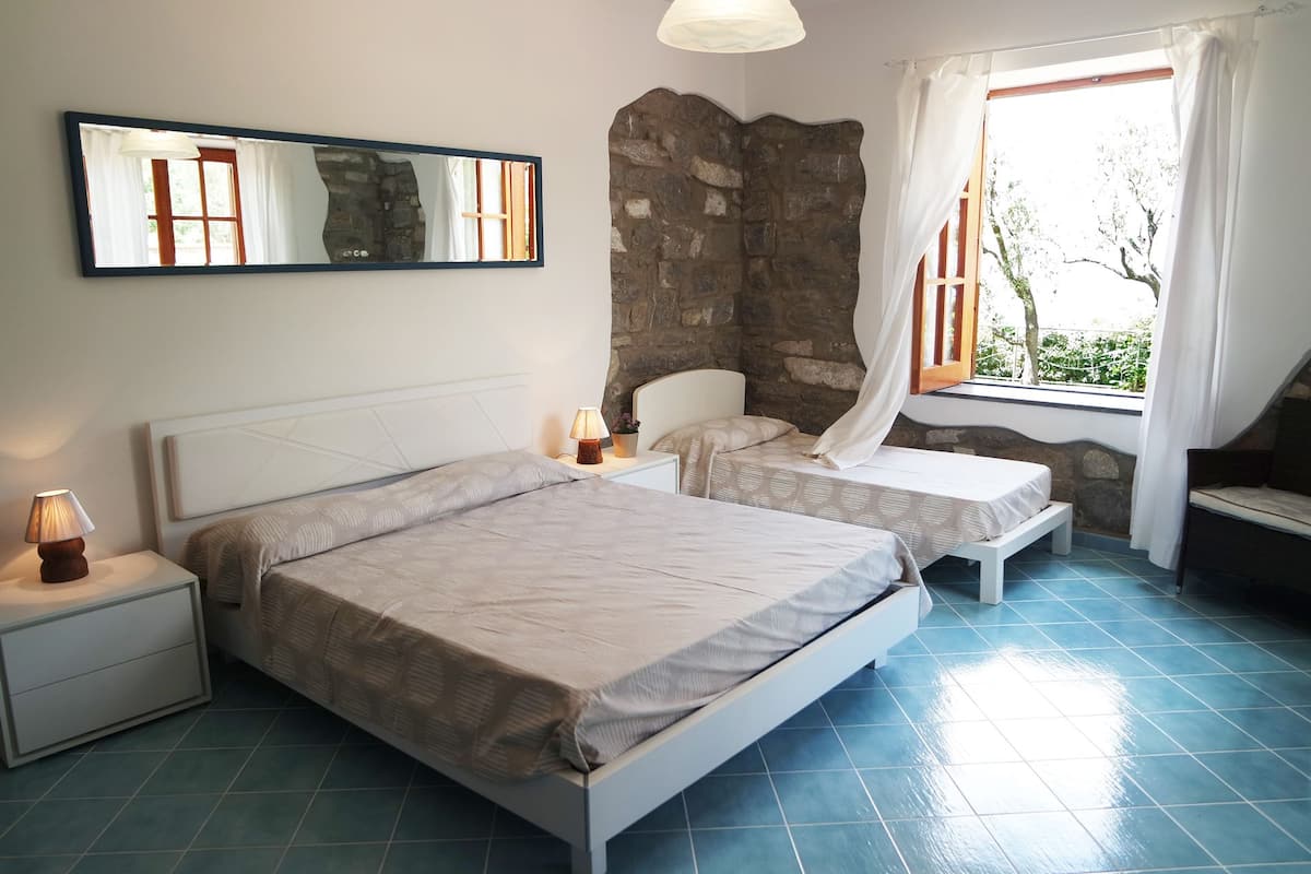 Un dormitorio con dos camas, una pared de piedra, una ventana con cortinas y una mesita de noche con lámpara.
