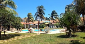 Área de piscina com espreguiçadeiras e guarda-sóis, cercada por palmeiras e um edifício com telhado telhado.