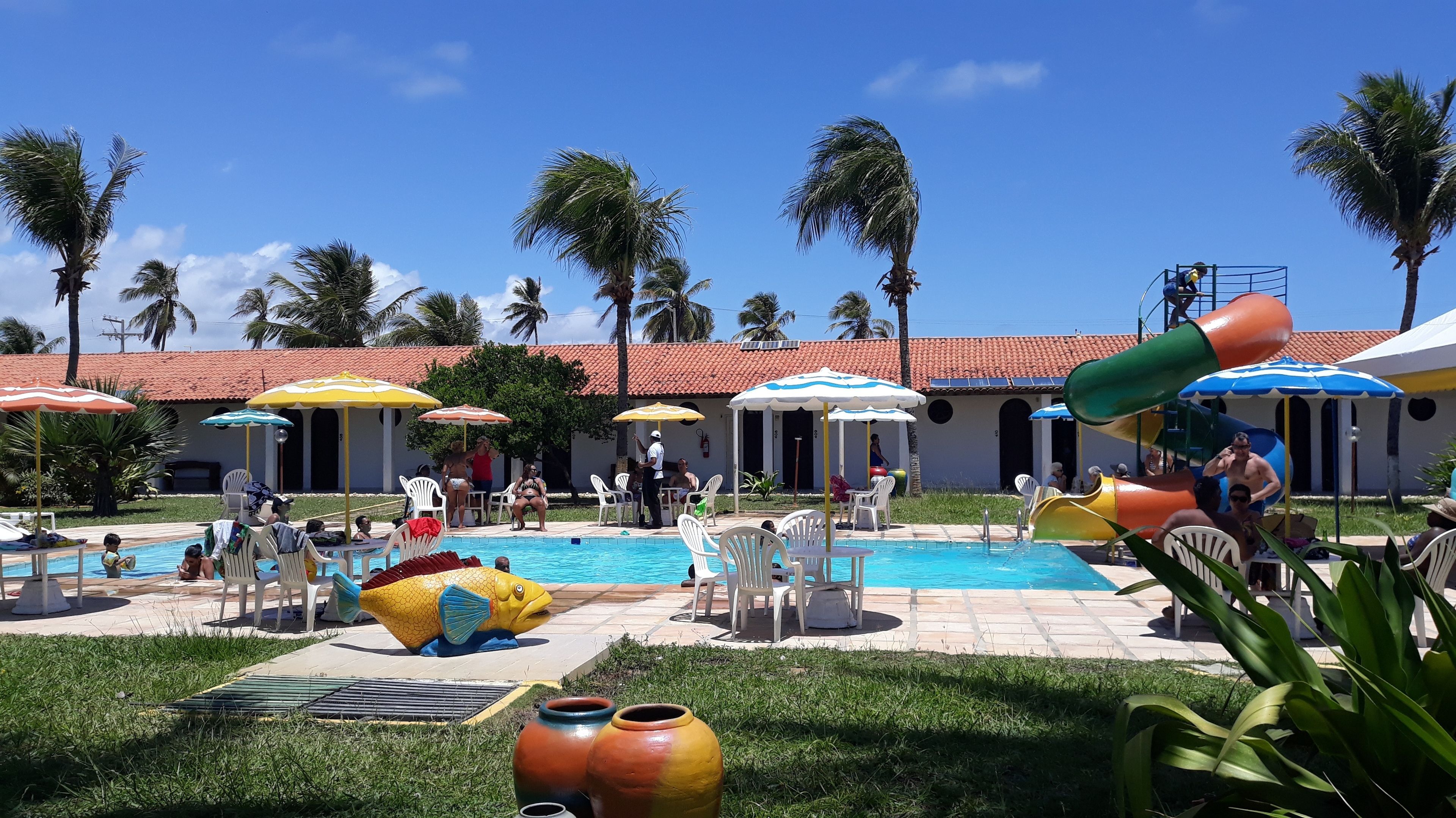 Uma área de piscina com vários guarda-sóis, uma torre de salva-vidas e palmeiras.