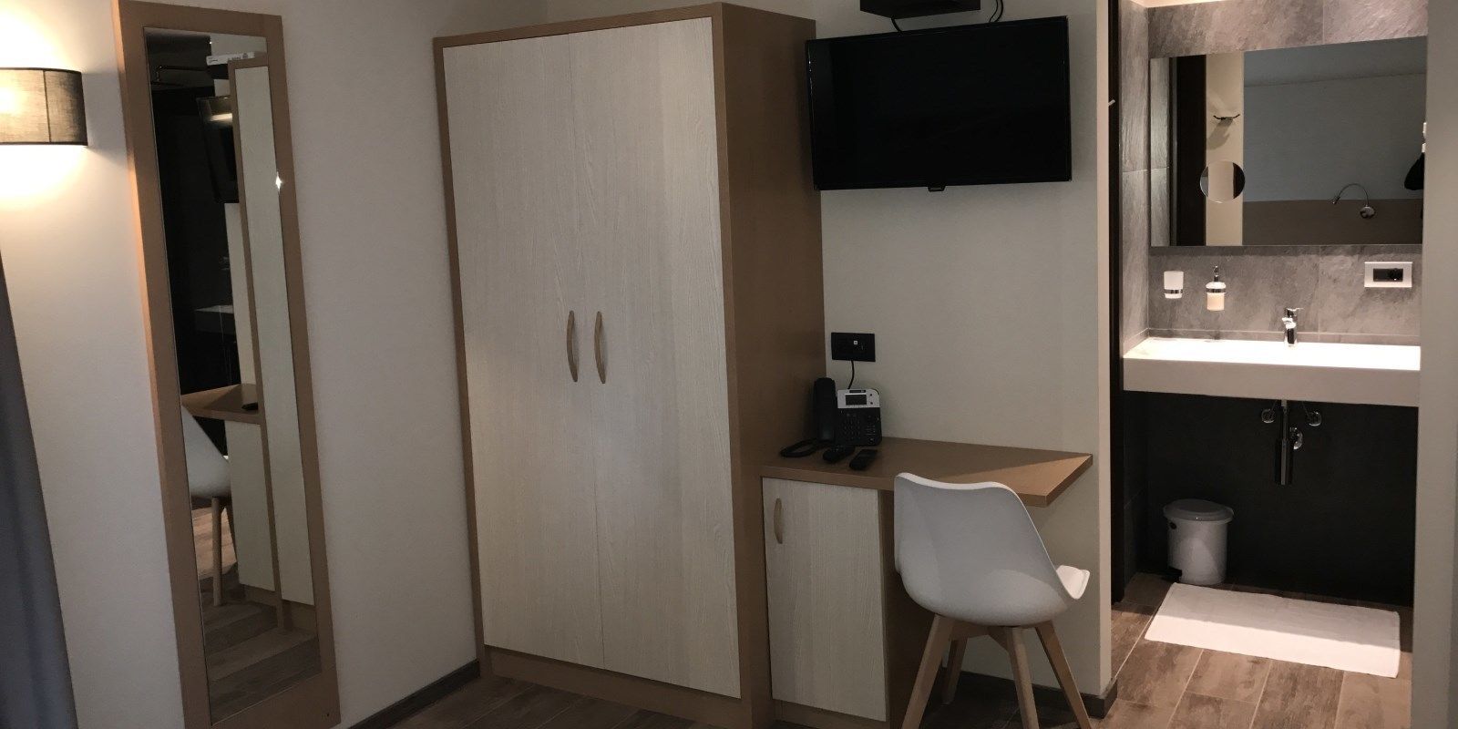 A compact room with a wardrobe, a desk with a chair, a flat-screen TV, a mirror, and a small bathroom with a sink and toilet.