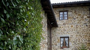A stone building with a small window and a door, surrounded by a stone wall and a grassy area with stone paving. - Caserio Iruaritz (Amurrio)