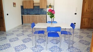 Un comedor con una mesa blanca y cuatro sillas azules, un florero con flores, y una cocina al fondo.
