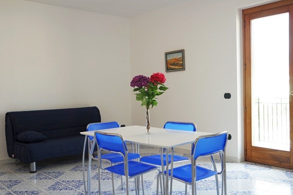A room with a white table, four blue chairs, a dark blue sofa, and a vase with flowers.