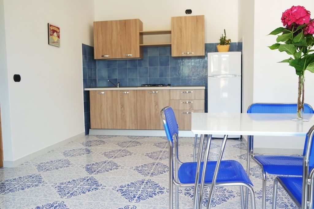 A modern kitchen with wooden cabinets, a white refrigerator, and a blue tiled backsplash. There is a dining table with blue chairs and a vase with flowers.
