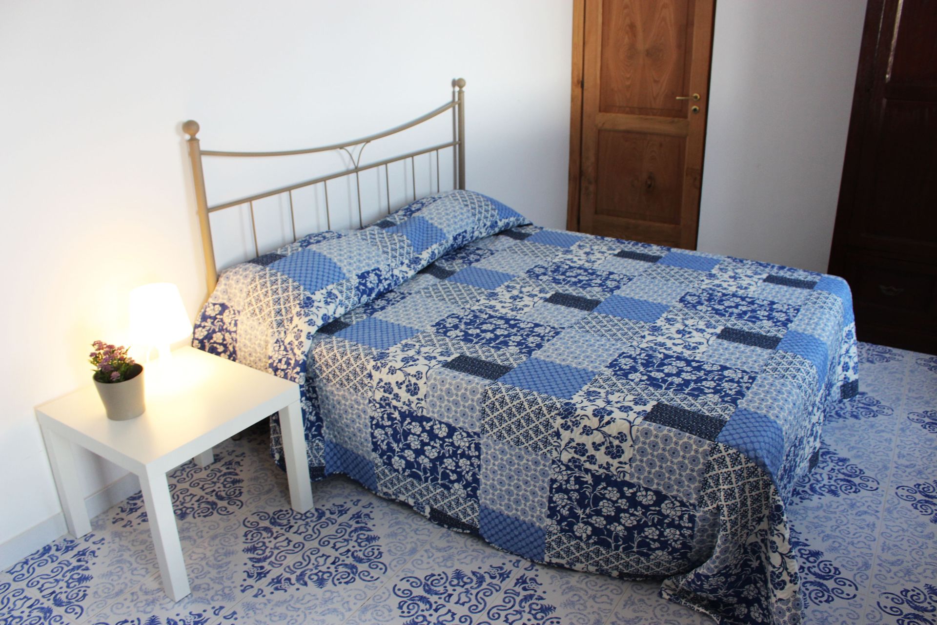 A bed with a blue and white quilt, a bedside table with a lamp, and a wooden door in the background.