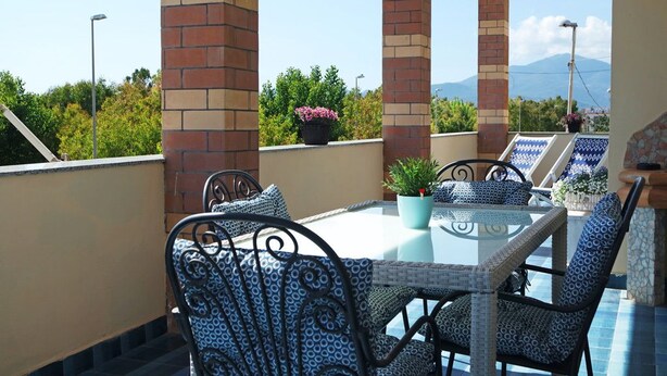 A rooftop terrace with a glass table and wicker chairs, surrounded by brick pillars and potted plants. - Casa Eleatica (Ascea)