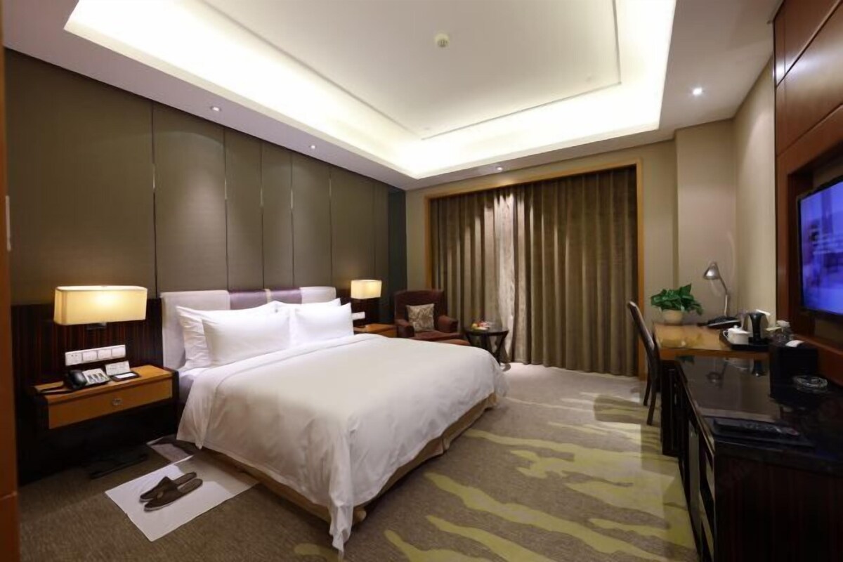 Habitación de hotel con una cama grande, un escritorio con ordenador, televisor y ventana con cortinas.