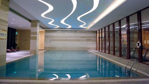 An indoor swimming pool with a modern design, featuring a clear pool, a wavy ceiling light fixture, and glass doors on one side. - Huifeng Hotel (Shuyang)