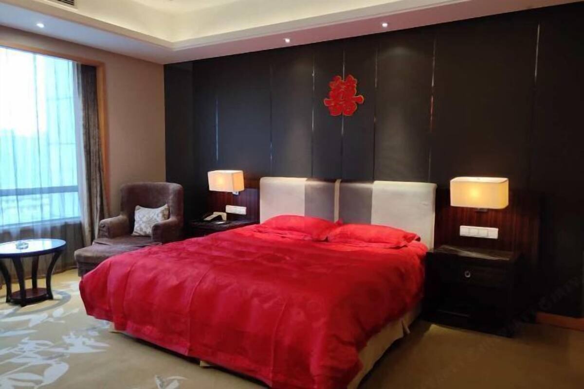 A hotel room with a large bed, two bedside tables with lamps, a chair, a small round table, and a large window with curtains.