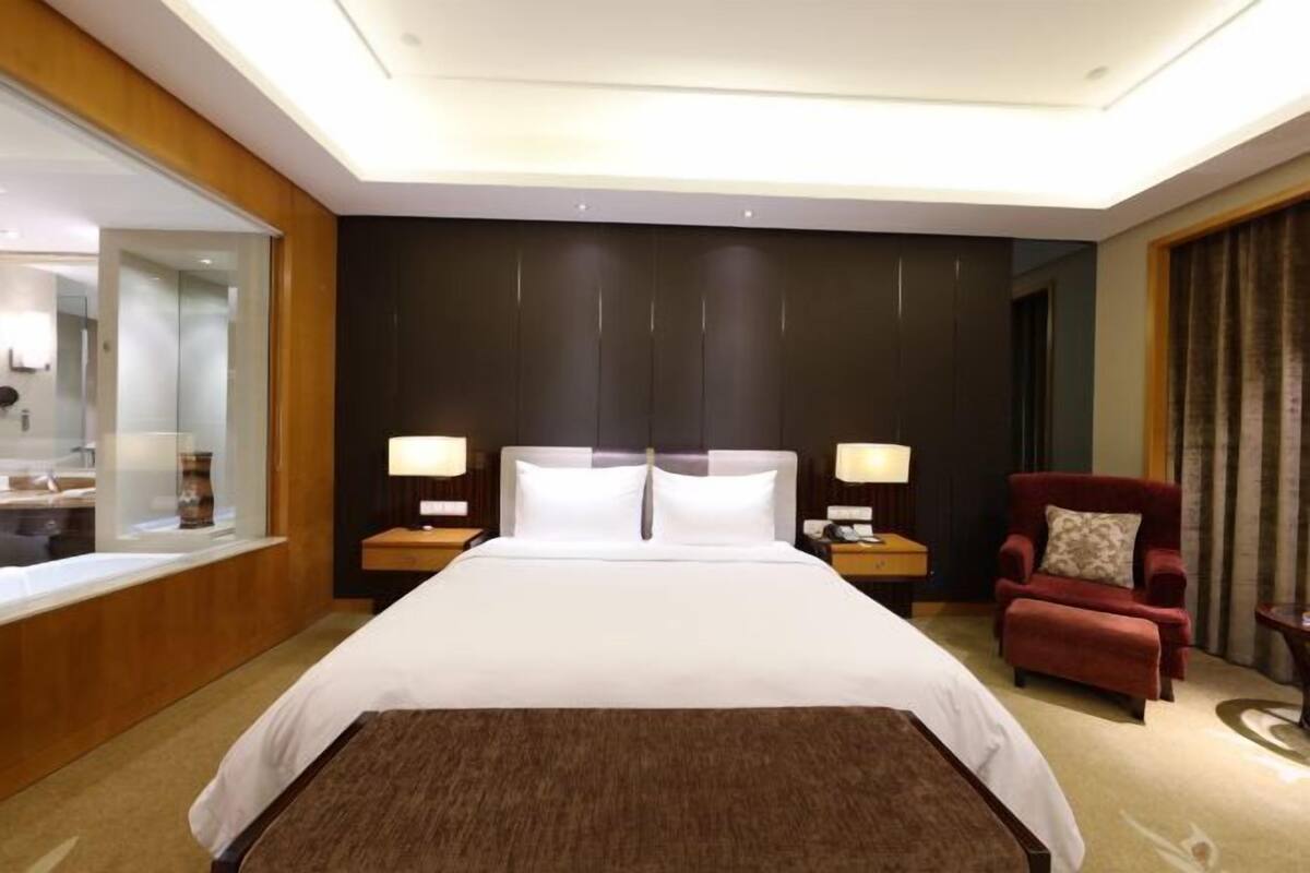 A hotel room with a large bed, two bedside tables with lamps, a red armchair, a wooden panel wall, and a large mirror.