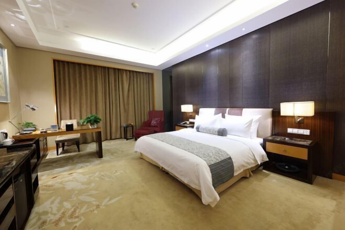Habitación de hotel con cama, mesitas de noche, escritorio, silla y un amplio ventanal con cortinas.