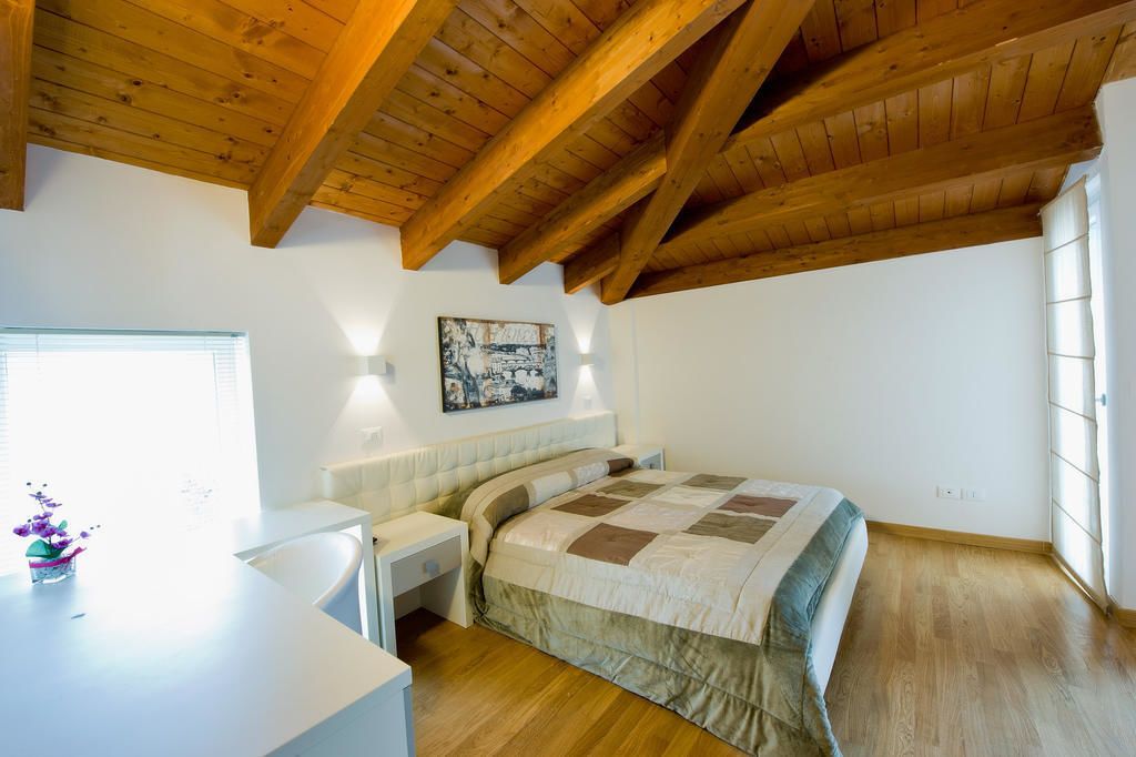 Une chambre avec un plafond en bois, un lit recouvert d’une courtepointe, une table de chevet, une commode, une fenêtre avec des stores et un luminaire fixé au mur.