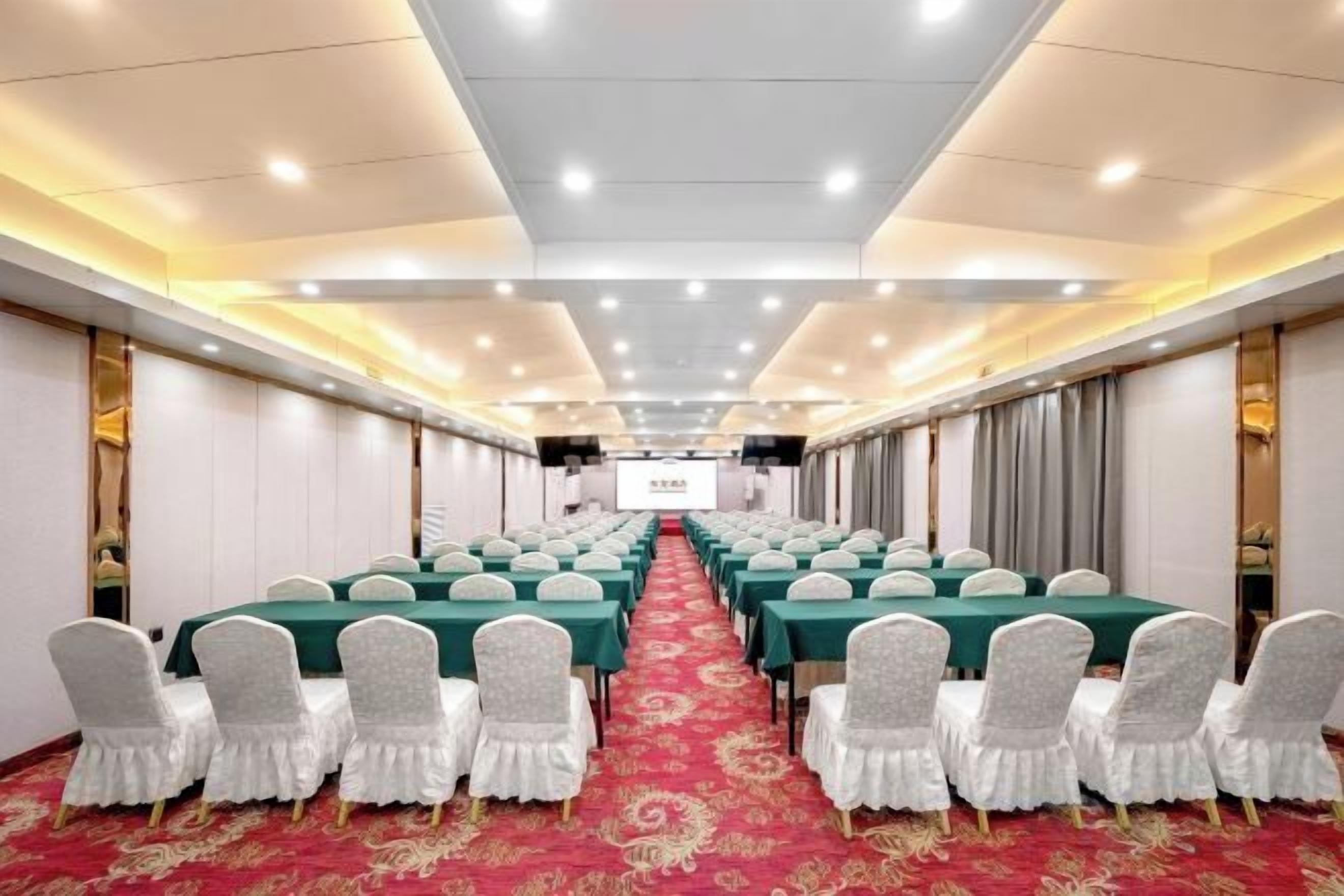 Un salón de conferencias con filas de mesas y sillas, una alfombra roja y una pantalla de proyector al frente.