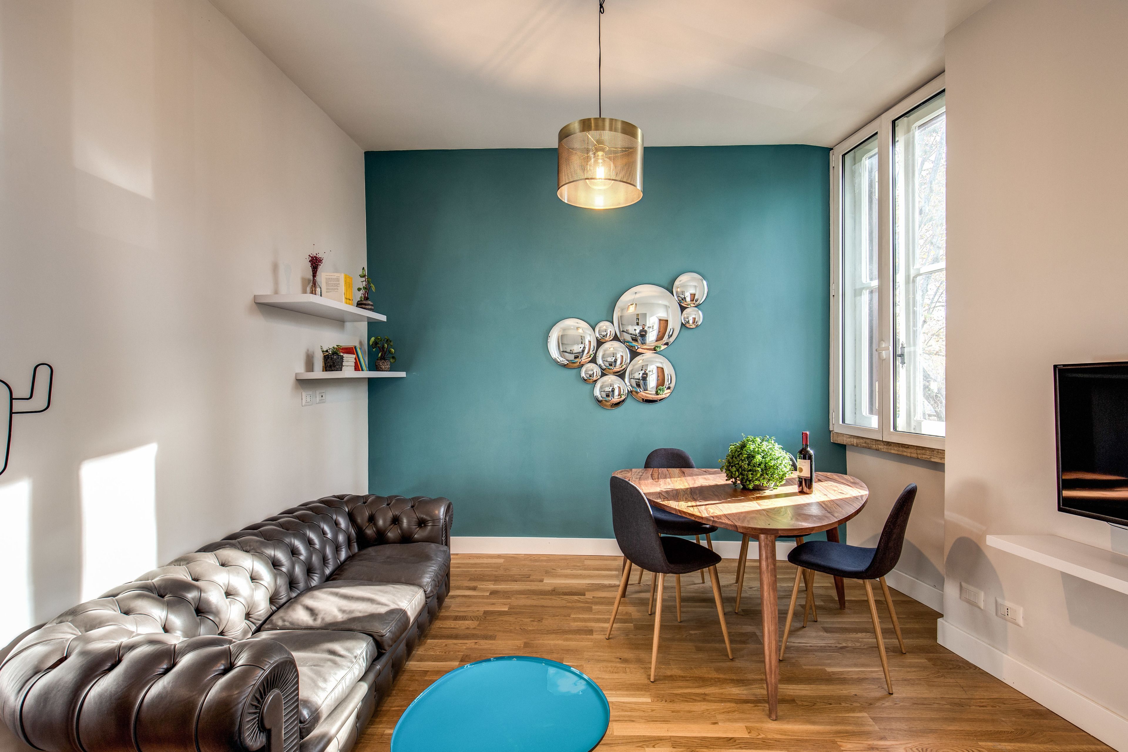 Una sala moderna con una pared de acento color turquesa, un sofá de cuero, una mesa de comedor con sillas y un televisor montado.