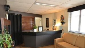 A reception area with a counter, a couch, a plant, and a window with blinds. - Auberge Happy Logis (Lac-Etchemin)