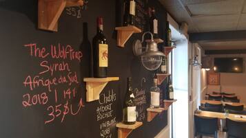 Interior de un restaurante con un menú en pizarra que enumera vinos y comidas, estantes de madera con botellas, y una ventana al fondo.