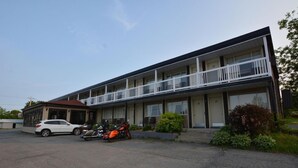 A motel with a two-story structure, multiple rooms, and a parking area with a white car and motorcycles. - Auberge Happy Logis (Lac-Etchemin)