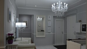 A hallway with a chandelier, a white sofa, a small table with a purple vase, and a door with a mirror and clock. - Ca dei Nobili (Genoa)