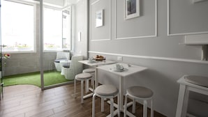 A modern, minimalist room with a glass door leading to a balcony with a green lawn. There are two white tables with stools, a small shelf with decorative items, and framed pictures on the wall. - Ca dei Nobili (Genoa)
